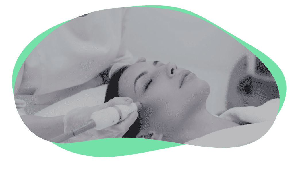 A woman lies on a treatment table with her eyes closed, receiving a facial treatment. A professional, wearing gloves, uses a handheld device on her cheek. The background is softly blurred, emphasizing the procedure.